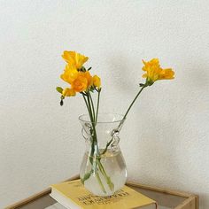 three yellow flowers in a clear vase on top of a stack of books next to a white wall