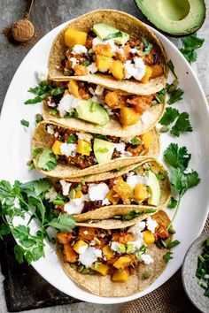 four tacos on a white plate with avocado and sour cream in the background