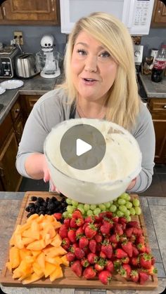 a woman holding up a bowl full of fruit on top of a wooden cutting board