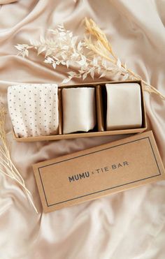 three square candles in a box on a white satin surface next to some dried plants