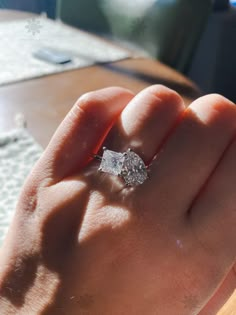 a person's hand holding an engagement ring on top of their finger, in front of a table