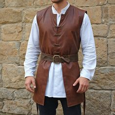 a man standing in front of a stone wall wearing a brown leather vest and white shirt