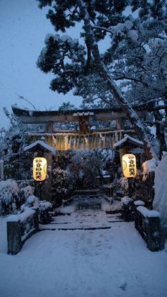 a snow covered path leading to a building with lanterns on it's sides and trees in the background