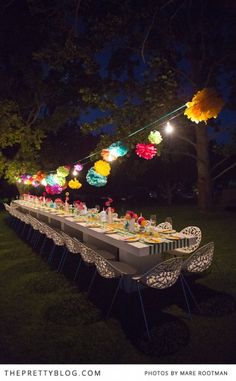 a long table is set up outside at night with colorful paper lanterns hanging from the trees