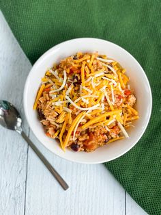 a white bowl filled with chili and cheese next to a spoon on a green napkin