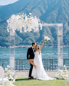 a newly married couple standing under an arch with flowers in front of the water and mountains