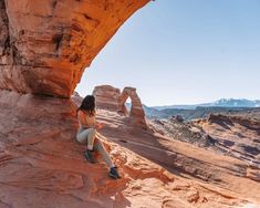 a woman sitting on the edge of a cliff looking at her cell phone in front of an arch