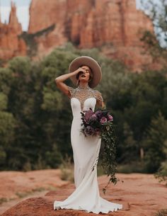 a woman in a wedding dress and hat standing on a rock formation with her hands behind her head