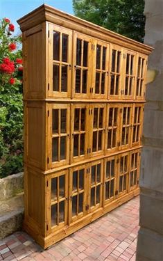 a large wooden cabinet sitting on top of a brick walkway next to flowers and bushes