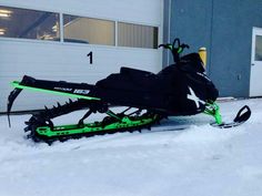 a green and black snowmobile parked in the snow next to a white garage door