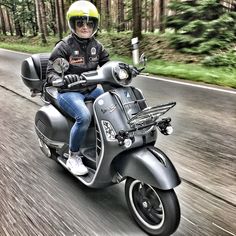 a man riding on the back of a silver motorcycle down a forest covered road with trees in the background