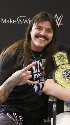 a man with long hair sitting in a chair holding a wrestling belt and pointing at the camera