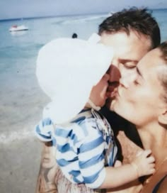 a man holding a baby on the beach with boats in the water behind him and his mother kissing him