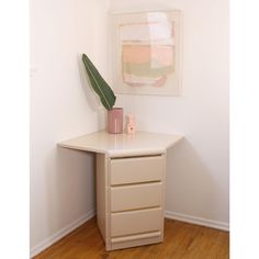 a white desk with a plant on top and a painting hanging above it in the corner