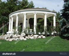 a white gazebo in the middle of a green lawn with rocks and trees around it