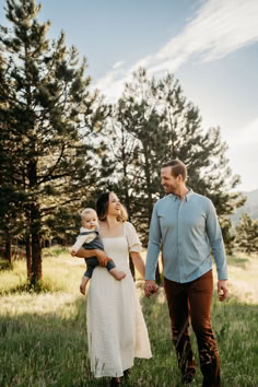 a man and woman holding a baby while walking through the grass with trees in the background