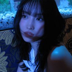 a woman with long black hair sitting in front of an aquarium looking at the camera