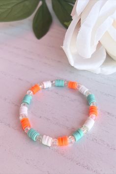 an orange and white beaded bracelet on a pink table next to a white flower