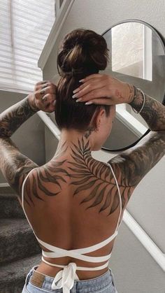 a woman with tattoos on her back standing in front of a mirror and looking at herself