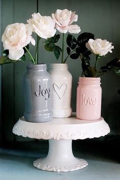 three jars with flowers in them sitting on a cake stand, one has the word love painted on it