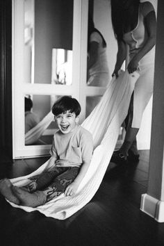 a young boy is sitting in a hammock and smiling at the camera while another woman looks on