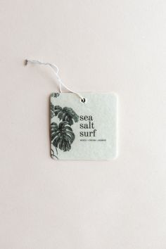 a sea salt and surf tag hanging from a string on a white wall with a plant