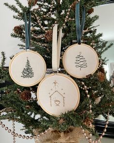 three cross stitch christmas ornaments hanging from a tree with pine cones and ribbons around them