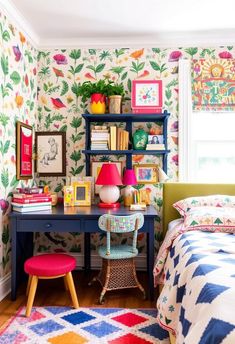 a bedroom with colorful wallpaper and a desk in front of the bed is filled with books