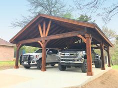 two trucks are parked under a covered structure in the driveway area with trees and grass behind them