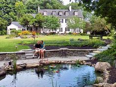 two people sitting on a bench next to a small pond in front of a large house