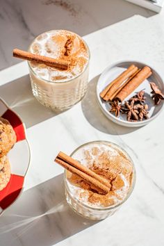 two glasses filled with drinks and cinnamon sticks on top of a white marble countertop