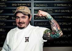 a man with tattoos and a baseball cap posing in front of a wall full of writing