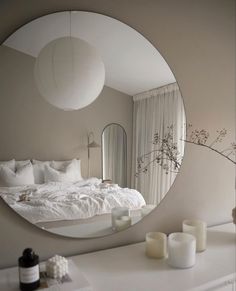 a white bed sitting under a round mirror next to a table with a vase on it