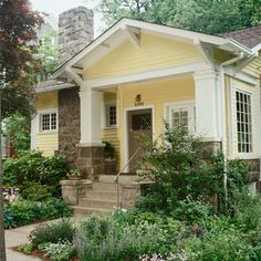 a small yellow and white house with trees in the front yard, bushes on either side