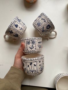 a person holding four cups in front of several other mugs on a white surface