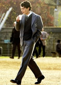 a young man in a suit and tie walking across a field holding a coffee cup