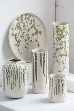 four white vases with green designs on them sitting on a shelf next to plates