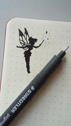 a black marker pen sitting on top of a white notebook with a drawing of a fairy