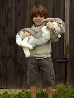 a young boy holding a cat in his arms and smiling at the camera while standing next to a wooden fence