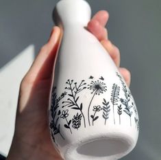 a hand holding a white vase with black flowers painted on the outside and inside it