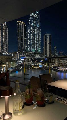 a view of a city at night from a restaurant table with drinks and glasses on it