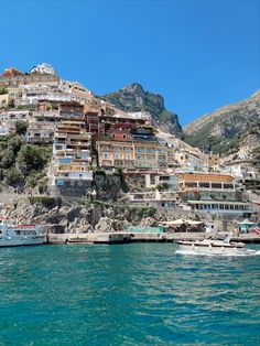 boats are parked on the water in front of a hillside with buildings and mountains behind it