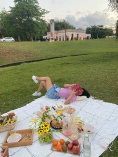 two women laying on a blanket in the grass with food and drinks laid out around them