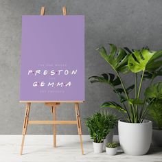 an easel with a purple poster on it next to potted plants and a plant