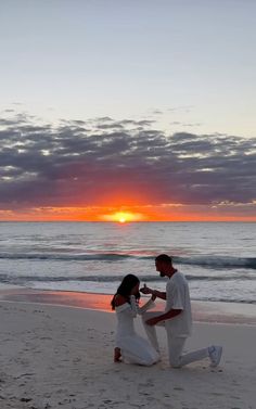 a man kneeling down next to a woman on top of a beach near the ocean