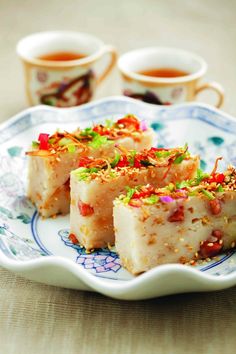 three pieces of cake on a plate with two cups of tea in the back ground