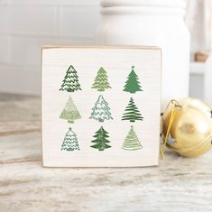 a wooden block with christmas trees on it next to ornaments and a gold ornament