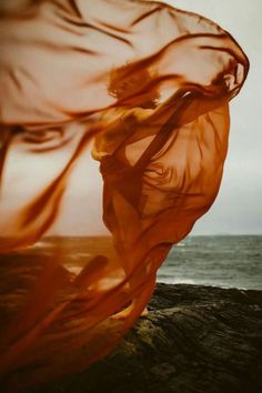 a woman is standing on the rocks by the ocean with an orange flowing fabric in front of her