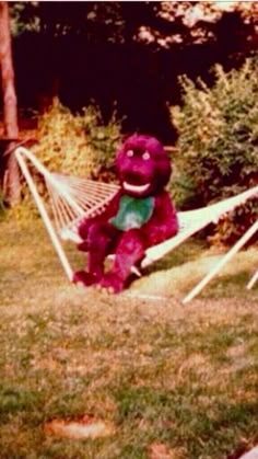 a purple teddy bear sitting in a hammock on the grass next to trees