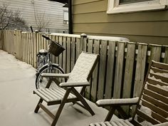 two lawn chairs and a bicycle are covered in snow near a fence with a wooden slatted fence